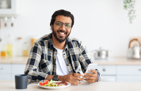 Cheerful middle-eastern young man with long hair and stylish beard in casual enjoying breakfast, drinking coffee, holding smartphone, indian guy freelancer checking emails in the morning, copy space