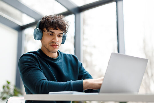 Concentrated young european man wearing headphones is intently using laptop while seated at modern workspace with large window in the background