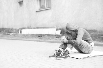 Homeless woman with paper cup on carton outdoors, space for text. Black and white effect