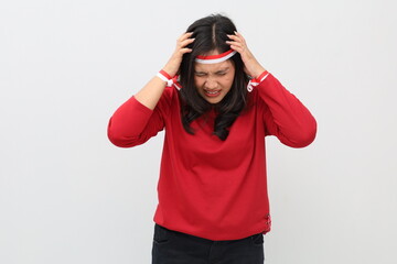 Frustrated Indonesian woman with red and white ribbon on head celebrating Indonesian independence day, standing holding head and covering ears, isolated on white background.