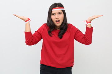 Indonesian woman screaming in confusion with red and white ribbon, celebrating Indonesian independence day standing while making question gesture, raising open hands, isolated on white background.