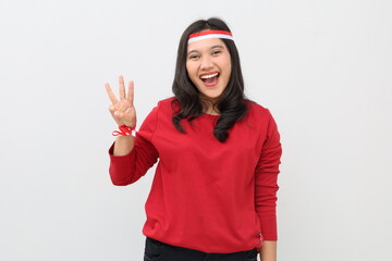 Indonesian woman with red and white ribbon, celebrating Indonesian independence day, smiling looking at camera, standing while showing three fingers, number three.