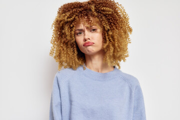 Sad girl with curly hair in a blue sweater, expressing disappointment, against a plain white...