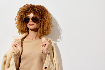 Stylish woman with curly hair, wearing sunglasses and a beige sweater against a plain white...