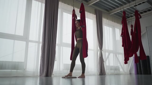 Lower angle of aerial yogi gripping red silk fabric overhead in bright studio, mindful balancing on two wooden nail boards with one foot on each for reflexology and proprioceptive training