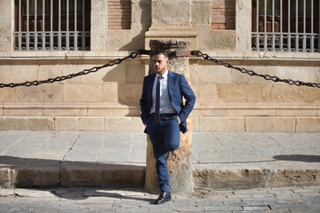 A young, handsome, dark-haired businessman wearing a suit, tie and black shoes waits impatiently with his hands in his pockets, leaning against a chained column in Seville's old town.