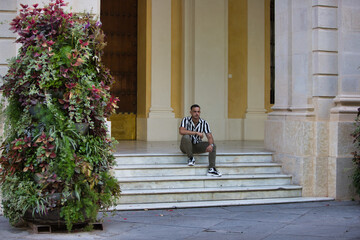 A young, handsome man with a beard, wearing a striped shirt and cargo trousers, is sitting on a white marble staircase in a building. The man is looking around.