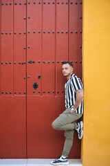 A young, handsome man with a beard, wearing a striped shirt and cargo trousers, rests with one leg on the doorframe of a house with a red Mediterranean-style door in Seville, Spain.