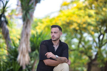 A young, handsome gay man with a beard, tattoos, earrings and modern clothes is in the park, resting his arms on his leg, which is propped up on a bench, and looking seriously to the side.