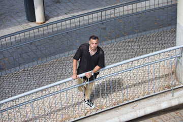 A young, handsome man with a beard, wearing a black shirt and beige cargo trousers, is holding onto...
