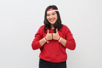 Smiling Indonesian woman standing celebrating Indonesian independence day with red and white ribbon, showing thumbs up, appreciating something, isolated on white background.
