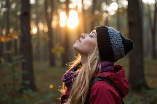 A woman with a beanie and backpack gazes upwards in a serene forest, bathed in golden sunlight. - Powered by Adobe