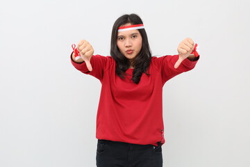 Indonesian woman unhappy with red and white ribbon celebrating Indonesian independence day while showing thumbs down sign of dislike, isolated on white background.