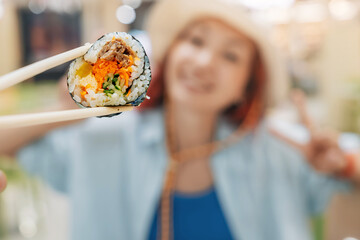 Chopsticks holding gimbap, a korean dish, with a smiling asian woman blurred in the background