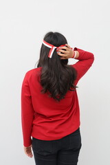 Rear view of a woman with a red and white ribbon celebrating Indonesian independence day while holding her head and waist, a gesture of thinking about something or being confused.