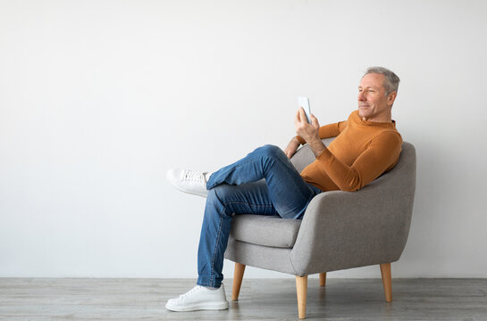 People And Technology Concept. Portrait of casual cool mature man resting and using smart phone sitting on chair isolated on white studio background wall, free copy space mockup, full body length