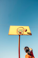 Unrecognizable teen girl throwing basketball toward hoop on outdoor court under clear blue sky, dynamic upward angle with ball nearing basket, youth sport, motivation, athletic, motion blur