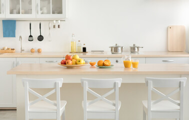 Vitamins, fruits, health care and stylish cuisine in new rent flat. Plate with apples and fruits, glasses with jug with juice on table, white chairs and utensils on furniture, light walls, copy space