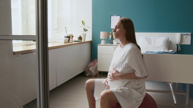 Medium full shot of heavily pregnant Caucasian woman bouncing on birthing ball, breathing through painful contractions, practicing deep breathing technique while preparing for labor in hospital