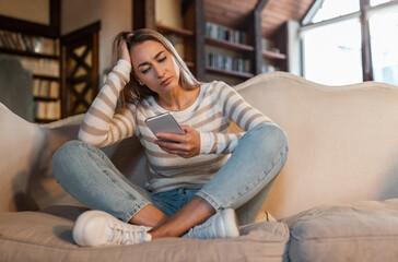 Worried lady looking at smartphone screen, sitting on couch at home, concerned middle aged woman...
