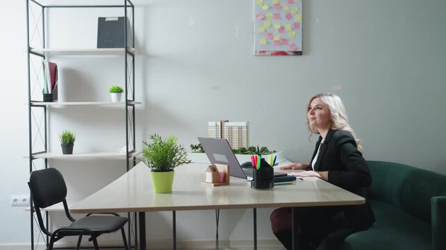 real estate agent seated in office welcomes guest with warm smile and gesture toward nearby chair, modern workspace setup with desk accessories, architectural model, laptop and green decor