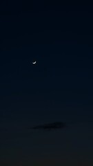 A slender crescent moon glows softly in the twilight sky above a faint cloud, evoking a calm and serene evening atmosphere.