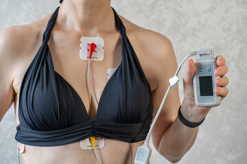 A young woman is demonstrating the use of a Holter monitor attached to her chest. The device is...