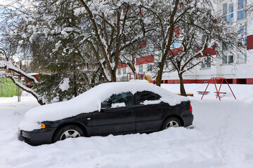 Snow-covered black car parked near residential building in winter. Urban courtyard with playground, leafless trees, and thick snow layer.