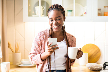 Portrait Of Happy Black Lady Drinking Morning Coffee And Using Smartphone In Kitchen At Home....