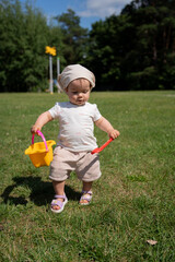 adorable toddler in sunlit park grasps bright yellow bucket and red spatula, enjoying a sunny day surrounded by green trees and a clear blue sky, capturing a moment of playful exploration, mood