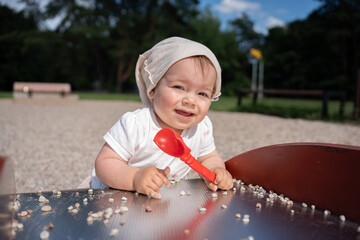smiling baby with red spoon enjoys a sunny day at the playground surrounded by tiny pebbles and...
