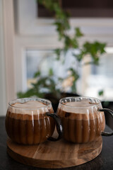 cozy indoor scene with two frothy cups of coffee on a wooden tray, surrounded by soft natural light filtering through a window with green plants in the background, inviting, luxury, refreshing