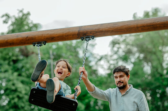 Cheerful boy swinging on swing with father standing in playground