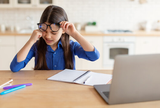 Eyestrain Problem. Tired Schoolgirl Rubbing Sore Eye Doing Homework On Laptop, Suffering From Fatigue And Dry Eyes Syndrome, Wearing Glasses Sitting At Desk At Home. Children Eyesight Issues