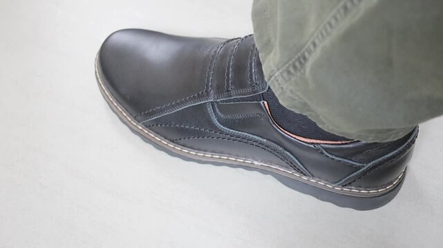 A man jerks and taps his foot in a black leather shoe on a gray floor close-up, 4K video