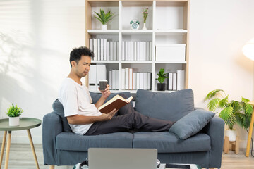 
A man is sitting on a couch reading a book while holding a cup. The room is well-lit and has a cozy atmosphere

