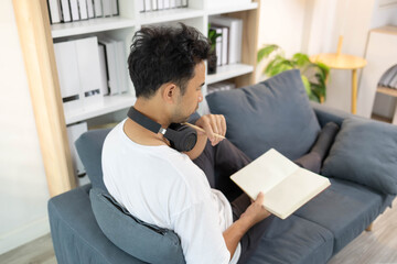 
A man is sitting on a couch reading a book while holding a cup. The room is well-lit and has a cozy atmosphere

