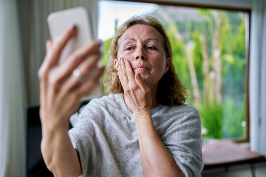 Senior woman looking at smart phone and touching face at home