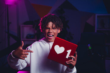 Young male content creator excitedly holding a heart symbol in a colorful indoor setup surrounded...