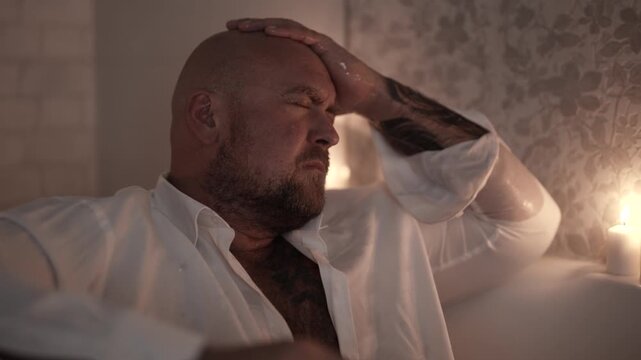 A man in a bathtub, in a white shirt, holding his head, a tense expression on his face.