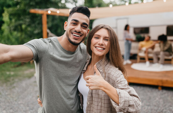 Loving young multiracial couple taking selfie near motorhome, resting with their friends outdoors. Arab guy and his Caucasian girlfriend enjoying summer camping trip, making mobile photo - Powered by Adobe