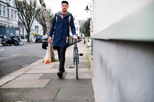 Young man wheeling with bicycle while holding bag of oranges on footpath at street