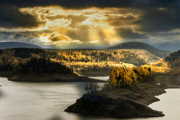 Dramatic sun rays pierce stormy skies over Hokkaido, illuminating golden autumn forests lining a serene lake. A breathtaking display of nature's power.