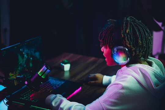 Teenager gamer using laptop and microphone under neon lighting during nighttime gaming or streaming session, wearing stylish hoodie