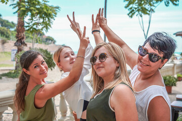 Four smiling women posing together on the beach raising their hands in a sign of peace, luck and friendship on a sunny day.