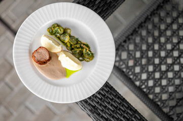 Gourmet dish with cooked meat, mashed potatoes, vegetables, and creamy sauce on a white plate, served outdoors on a woven chair. Top view of elegant food presentation.