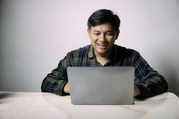 Happy young Asian man smiling broadly while using a laptop at a table, engaged in online activity...