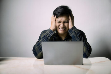 A young man clutches his head in extreme frustration and despair while looking at a laptop,...