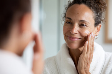 elegant woman in her 50s applying face cream in bathroom mirror, skincare and aging gracefully, natural window light