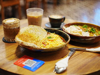 A set of Asian noodle dishes with papadam and coffee drinks served on a wooden table in a cozy café. Perfect for lunch or dinner vibes.